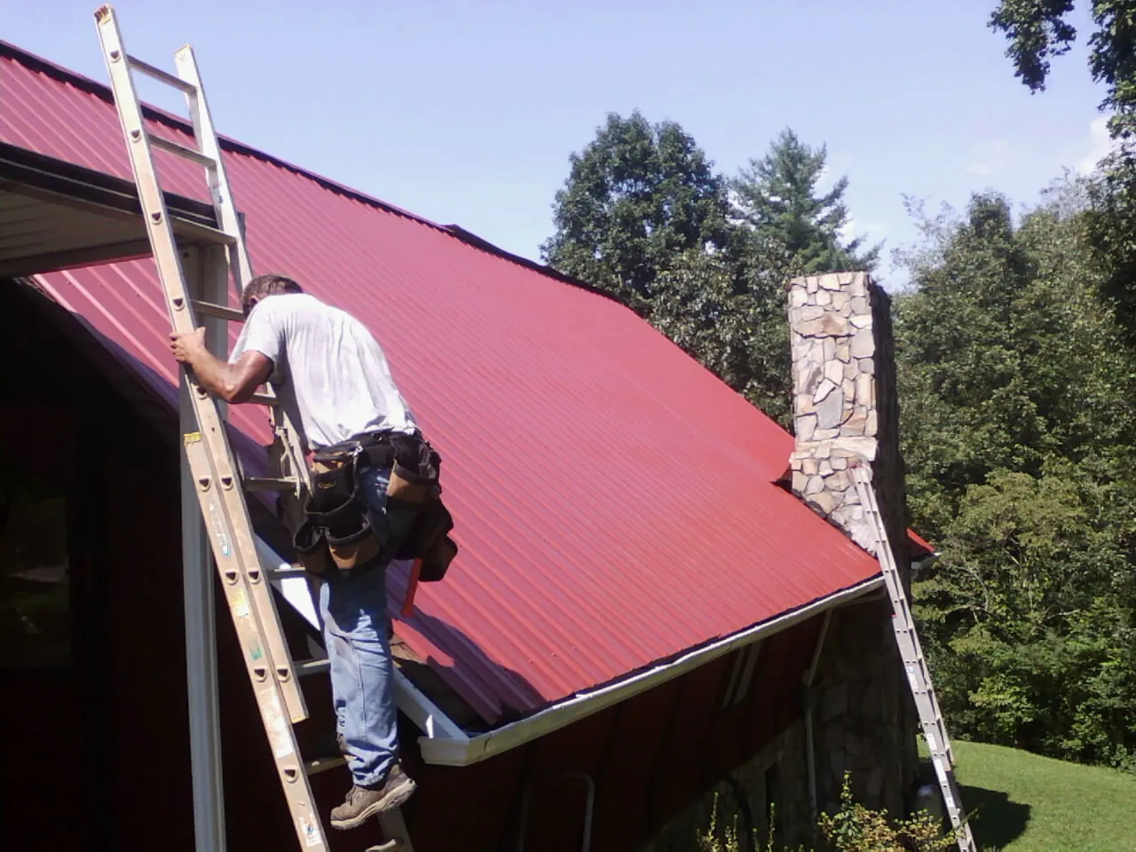 Roofer on a ladder beside a red metal roof and stone chimney — Soffit & Fascia Repair by Roofing Brothers in Denison